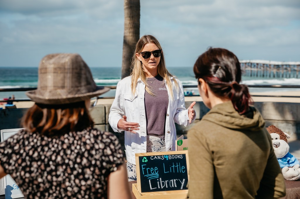 Trisha Goolsby speaking to visitors at an event on the beach boardwalk. A sign says cans4books and free little library.