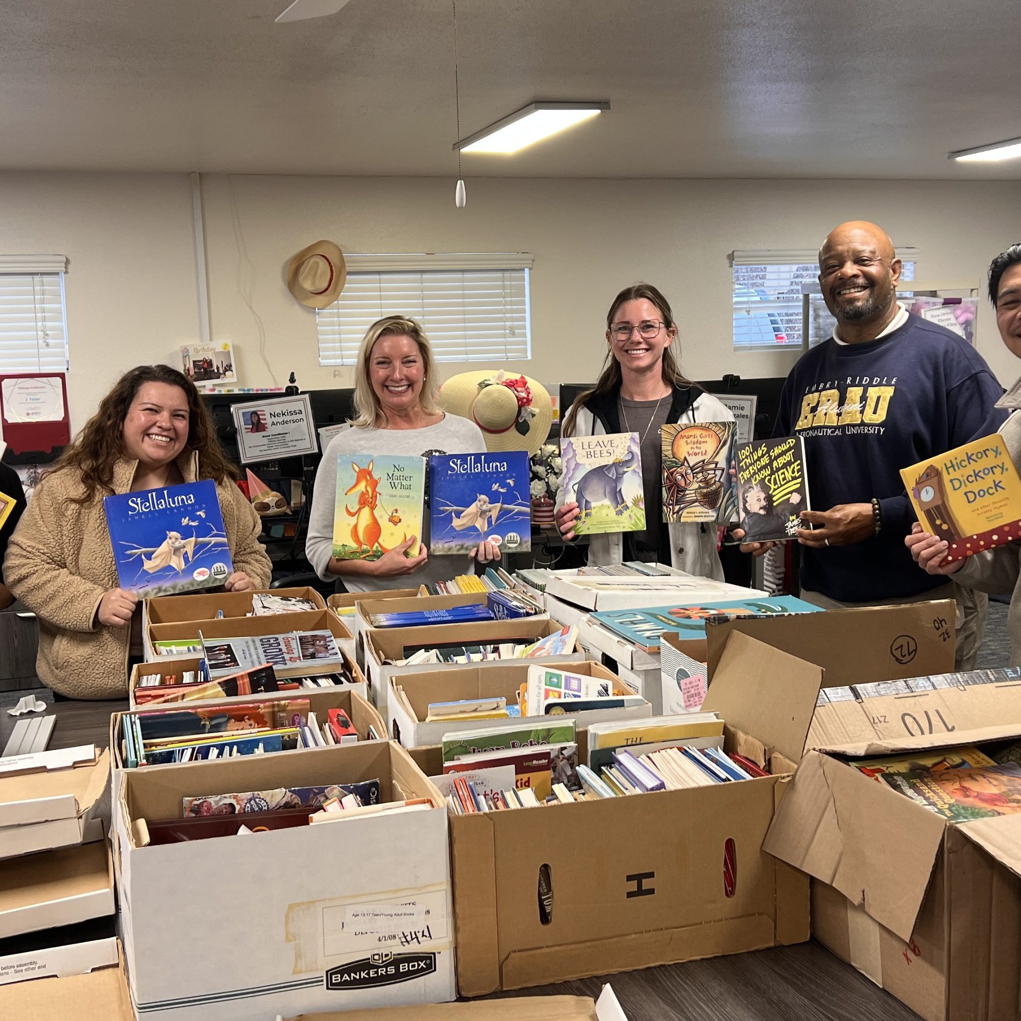 A group of people hold up children's books and smile. There are many boxes of books on the tables in front of them.